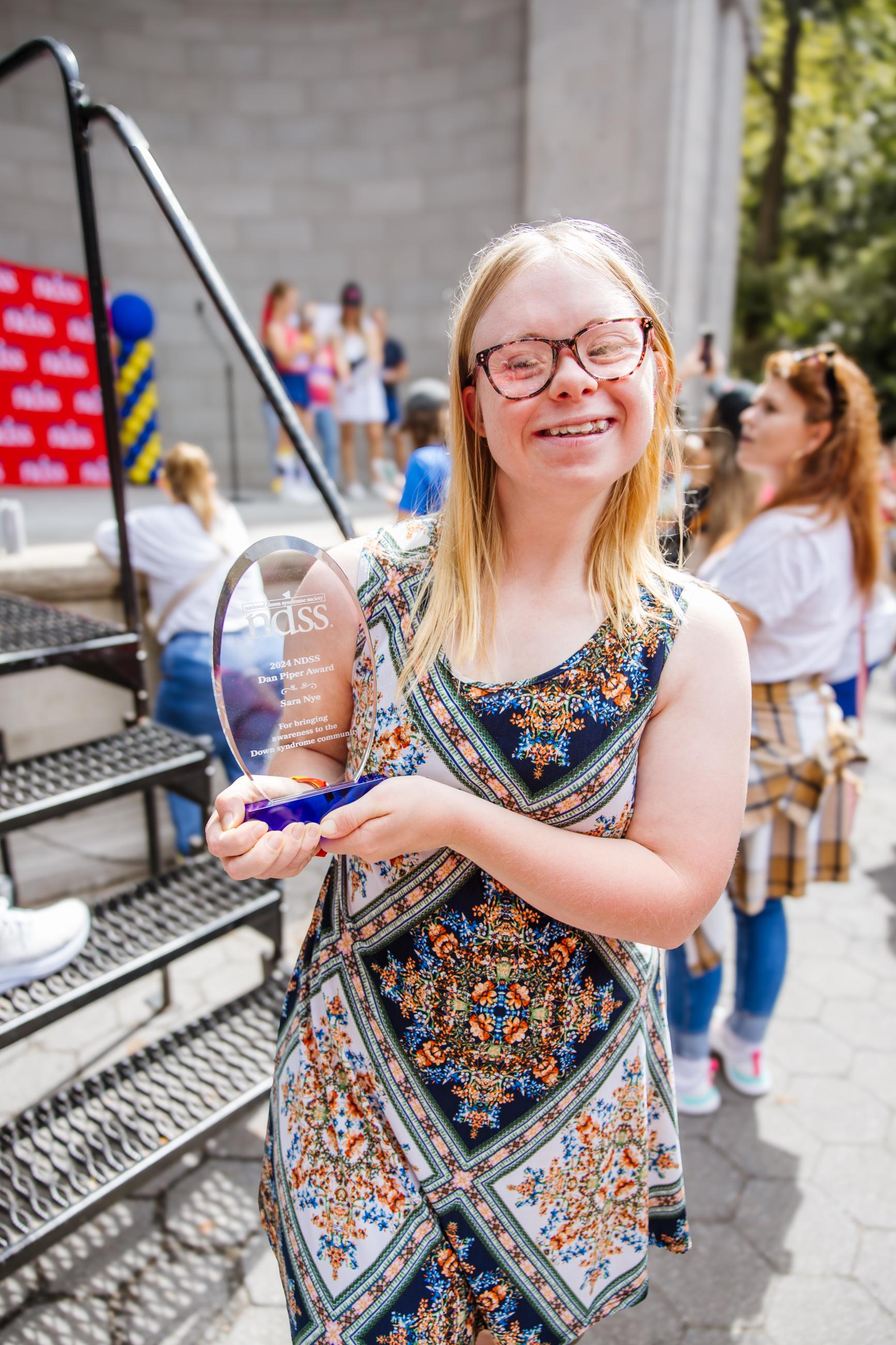 Woman with Down syndrome, blonde hair, and glasses smiles at the camera holding an award she won