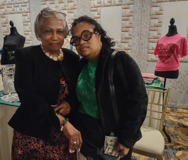 Black woman with Down syndrome wears black glasses, a green shirt, and a black sweater, stands in front of her business booth