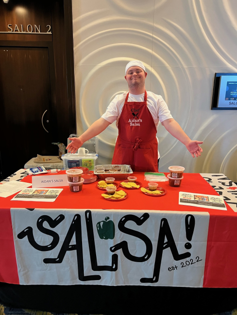 Aiden, a white man with Down syndrome, is wearing a white chef's hat and a red apron. He is gesturing to his red table with ingredients and jars. A large sign reads, "Salsa!"